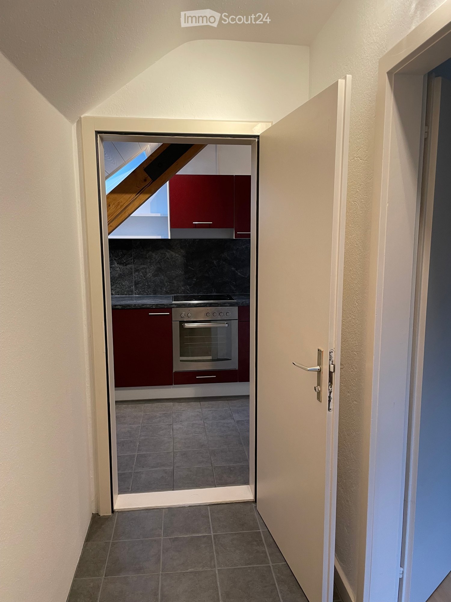 A narrow hallway with tiled flooring and white walls, leading to an open door that reveals a small kitchen area with red cabinets, a stainless steel oven, and a dark countertop.