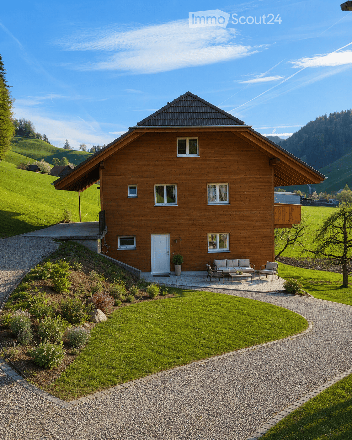 A two-story wooden house with a sloped roof, white door, multiple windows, and a small balcony. The house is surrounded by a well-maintained lawn, garden, and gravel driveway.