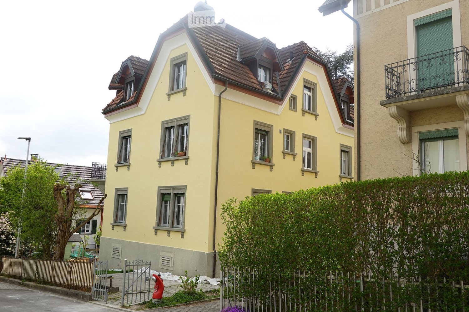 Two-story yellow house with multiple windows, dormer windows, and a red-tiled roof. The house is surrounded by greenery and a small fence.