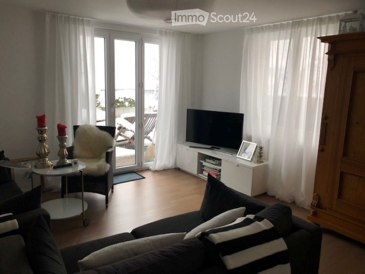 Modern living room with large windows, white curtains, dark wooden floor, sofa, coffee table, armchair, TV, and decorative items.