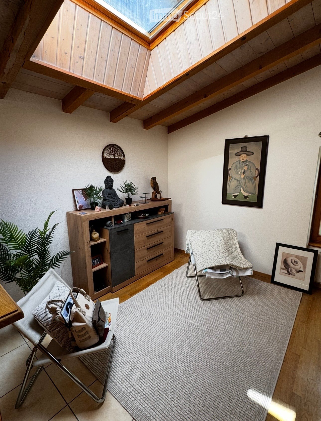 A cozy, well-furnished room with wooden floors, a skylight, a grey rug, a wooden cabinet with drawers, a chair with a blanket, and decorative items including a framed picture, a plant, and a small statue.