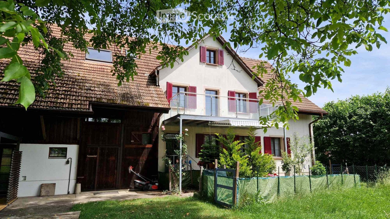 Two-story house with red shutters, tiled roof, balcony, and surrounding greenery.