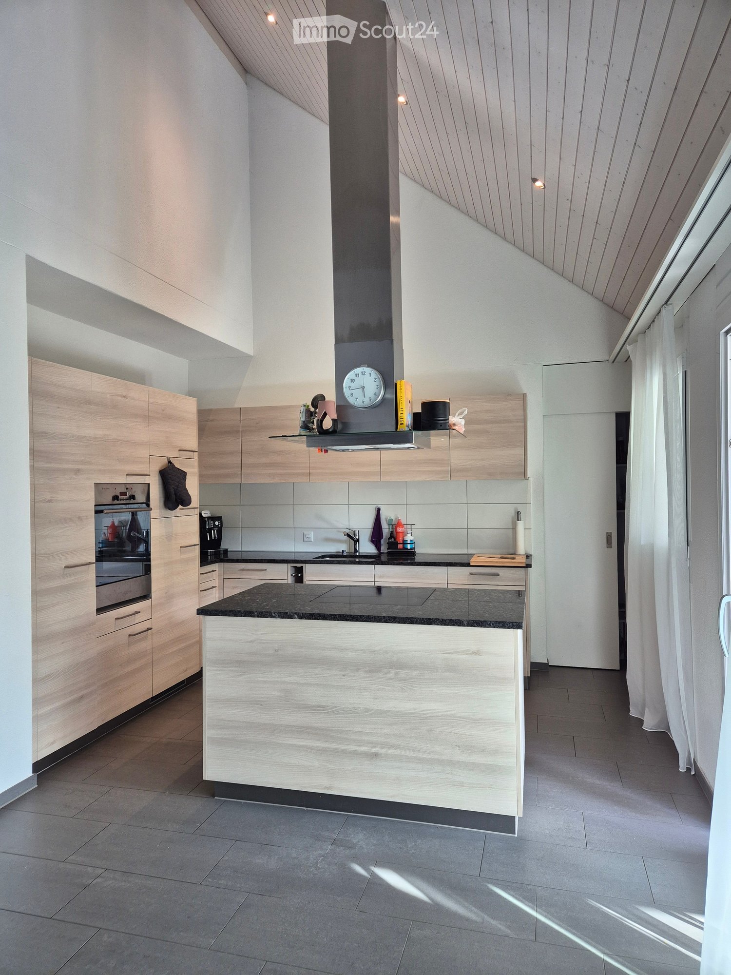 Modern kitchen with light wood cabinets, stainless steel appliances, and a central island with a black countertop. Features include a built-in oven, sink, and various small appliances.