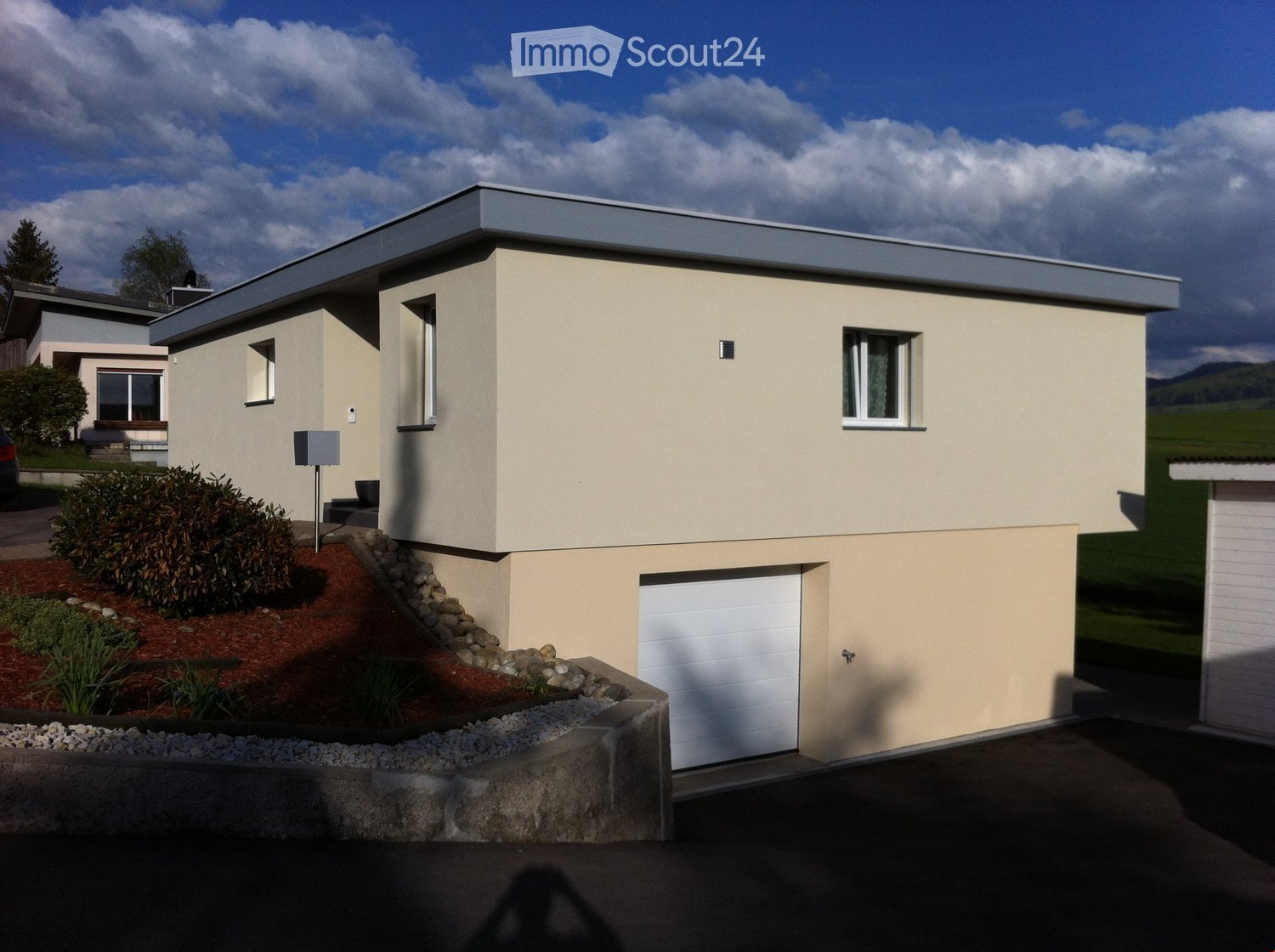 Single-story house with beige exterior, white garage door, and small garden area with shrubs and rocks.