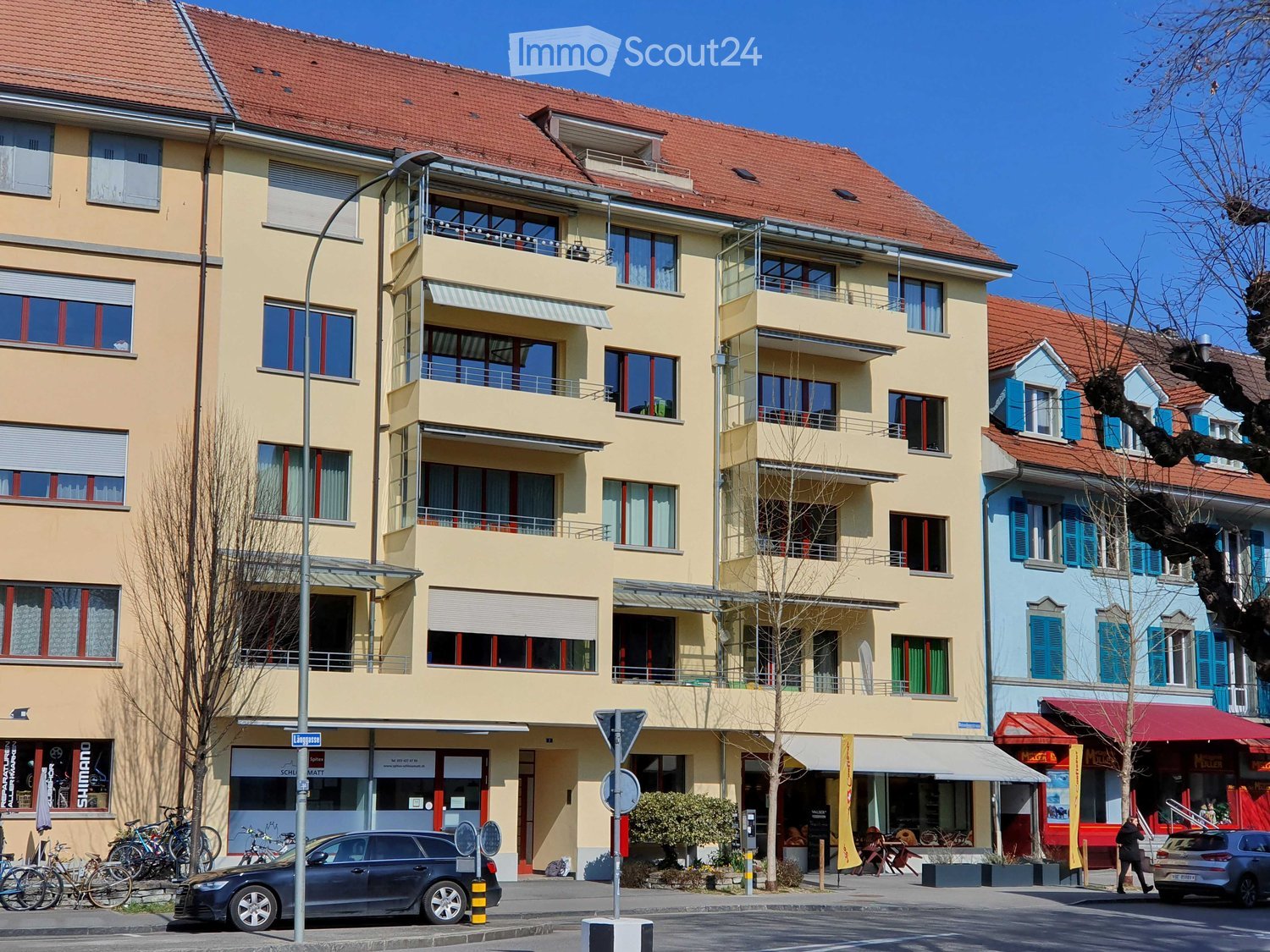Multi-story residential building with balconies, large windows, and a red-tiled roof. The ground floor features commercial storefronts with awnings and signage. Bicycles and a car are parked on the street.