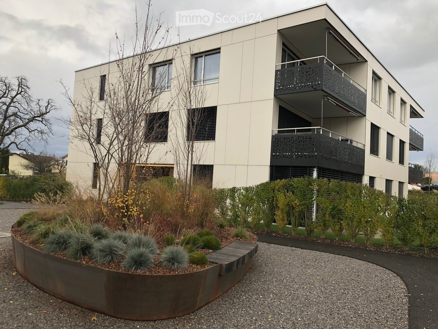 Building. Modern two-story building with balconies, surrounded by landscaped garden area with shrubs and a metal planter.