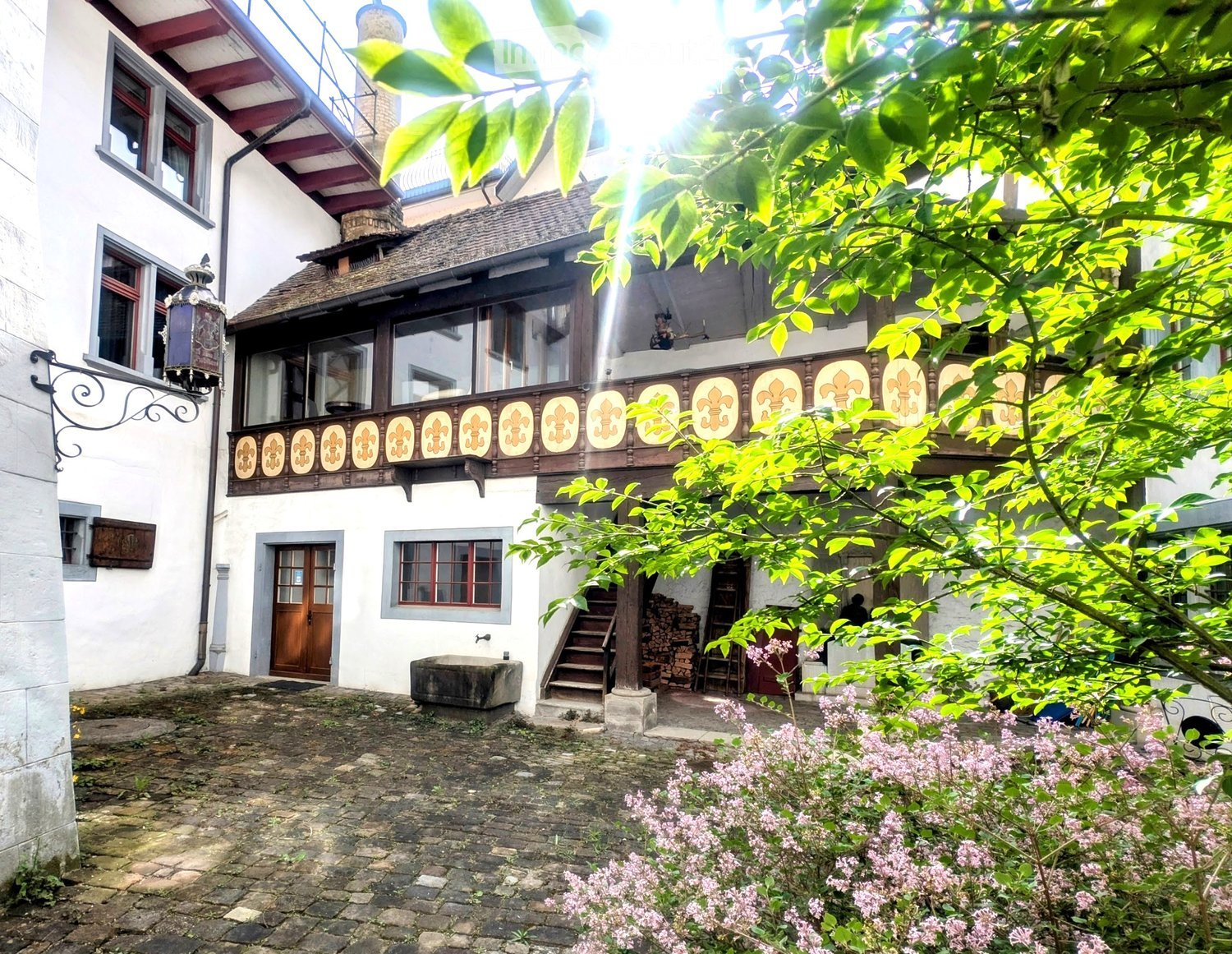A historic building with a balcony featuring ornate fleur-de-lis designs, surrounded by lush greenery and a stone-paved courtyard.