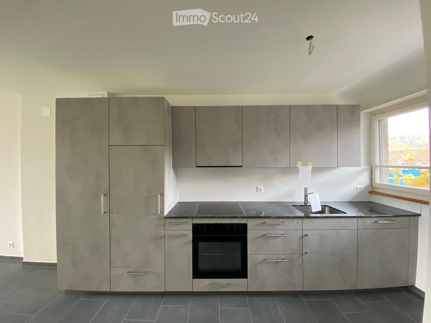 Modern kitchen with grey cabinets, stainless steel sink, and built-in oven. Features a large window with a view of the garden.