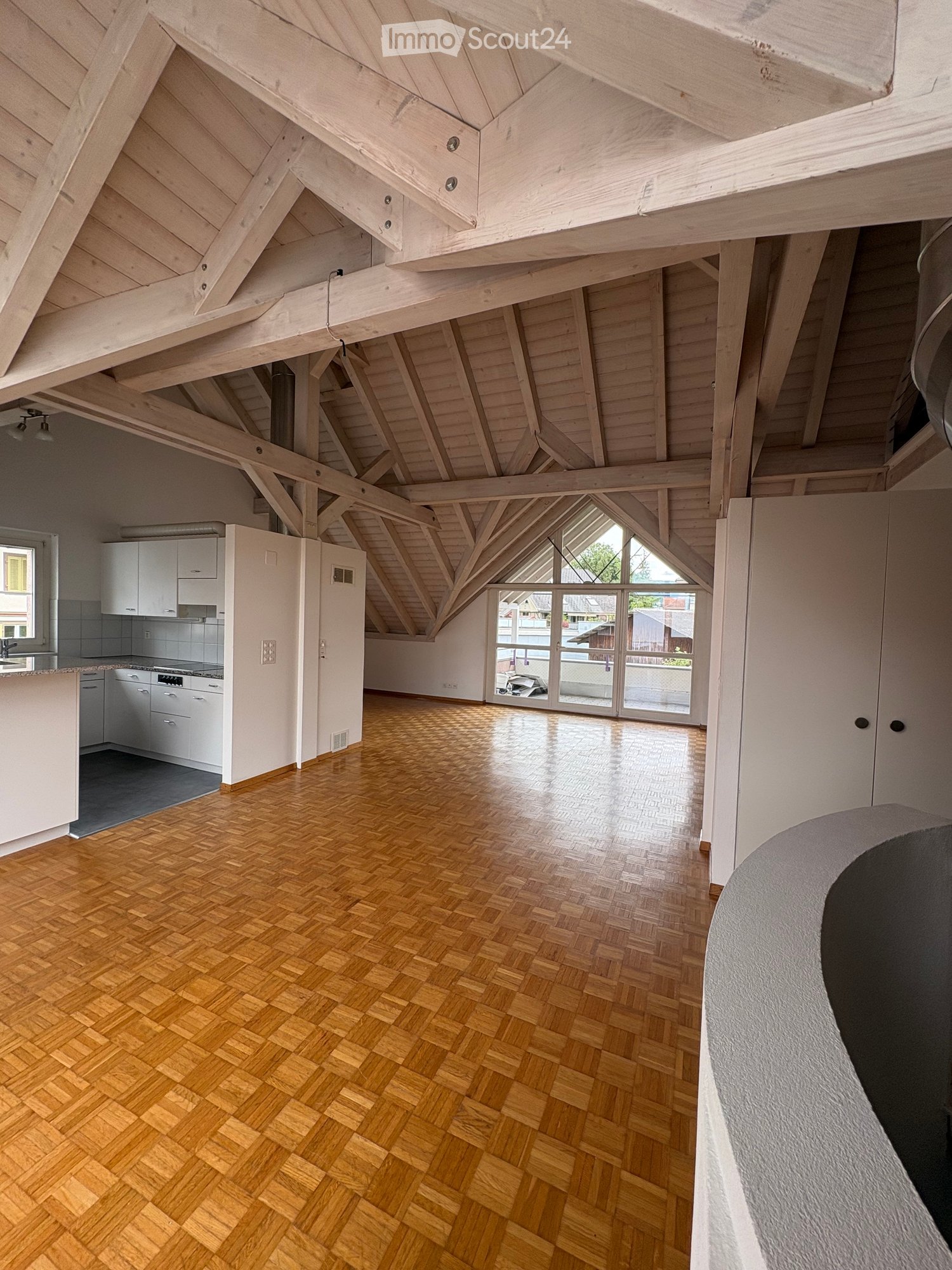 Open-plan attic space with exposed wooden beams, herringbone wooden flooring, and a modern kitchen area featuring white cabinets, a sink, and a stove.