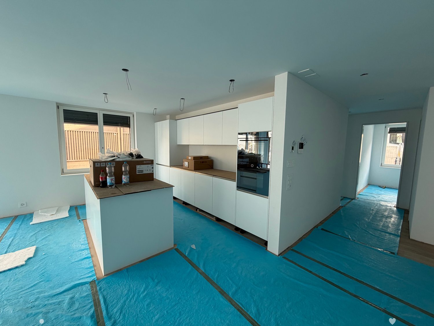 Empty kitchen with white cabinets, a countertop, and blue protective flooring. Windows provide natural light, and the space is in the process of being renovated.
