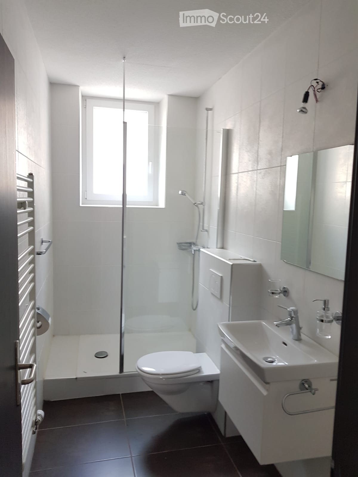 Modern, compact bathroom with a white toilet, sink, mirror, and glass-enclosed shower. Features a radiator on the left and a window above the shower area.