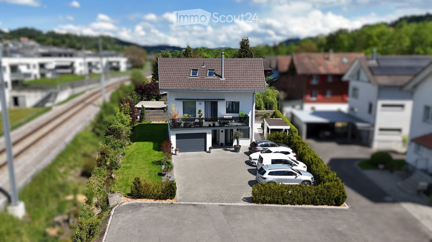 A modern two-story house with a tiled roof, garage, and balcony. The property features a well-maintained lawn, several parked cars, and surrounding greenery.