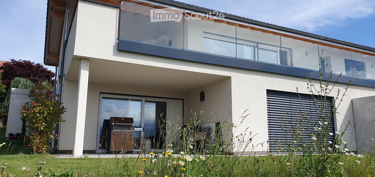Modern two-story house with a large glass balcony on the upper level, a covered patio area with a grill, and a well-maintained garden.