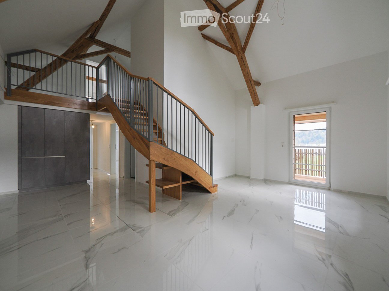 Spacious, modern hallway with polished marble floors, wooden staircase with metal railing, exposed wooden beams on the ceiling, and a large glass door leading to an outdoor area.