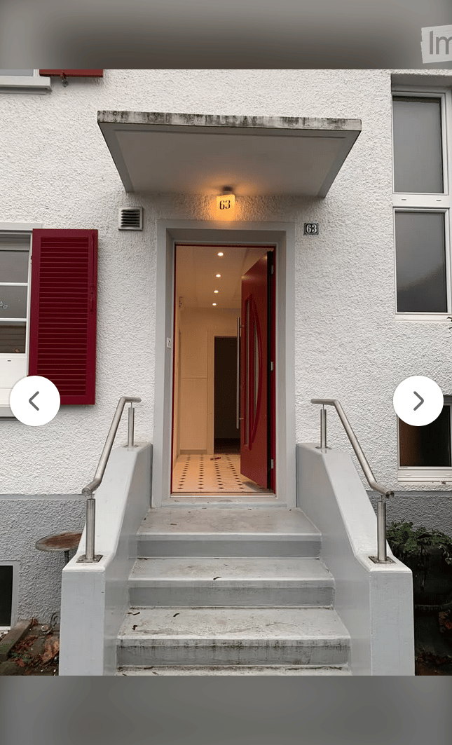 Exterior view of a two-story building with a raised ground floor, white facade, red shutters, and a small staircase leading to the entrance.