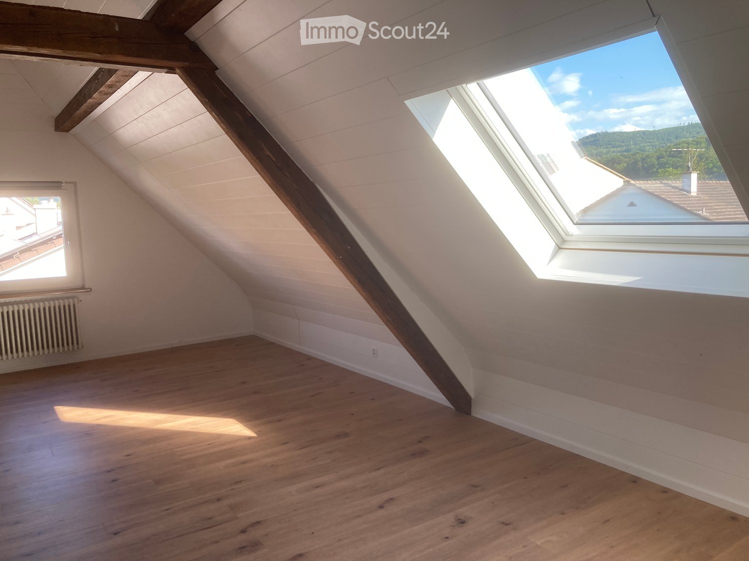 Empty attic room with wooden flooring, white walls, exposed wooden beams, skylight window, and a small window on the side.