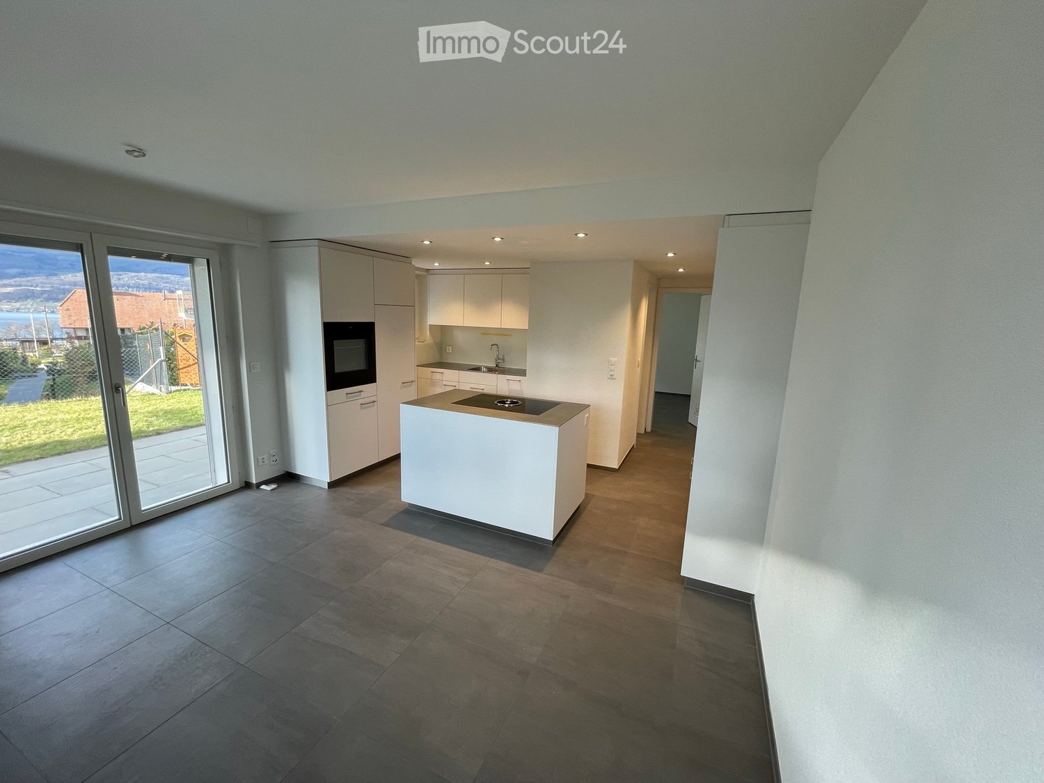 Modern kitchen with white cabinets, a central island, built-in oven, and a sink. The space features large windows and a view of the outdoor area.