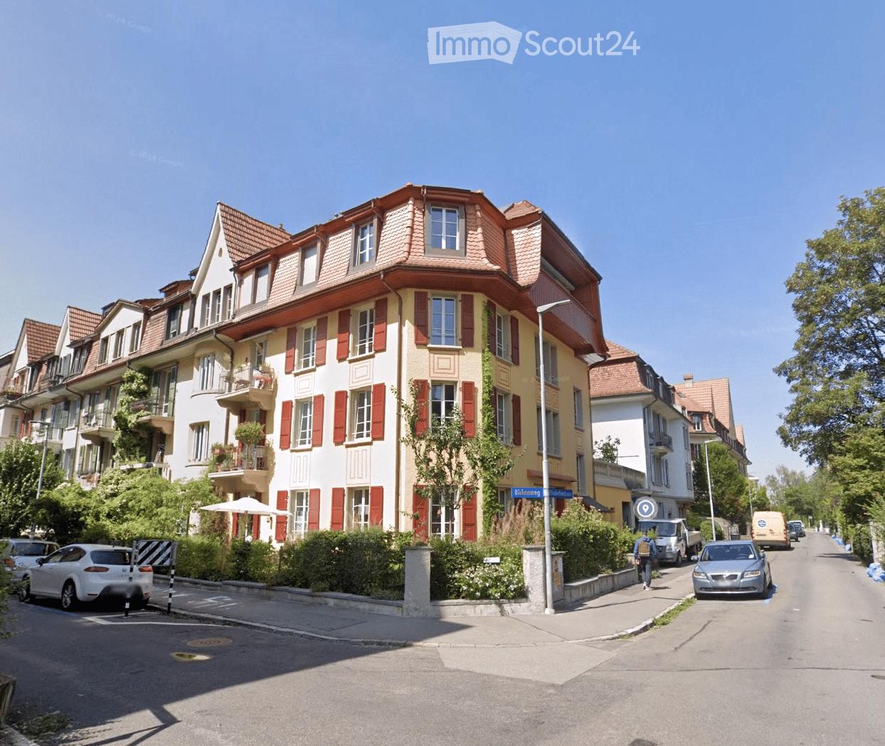Multi-story residential building with red shutters, balconies, and a red-tiled roof, situated on a street with parked cars and greenery.