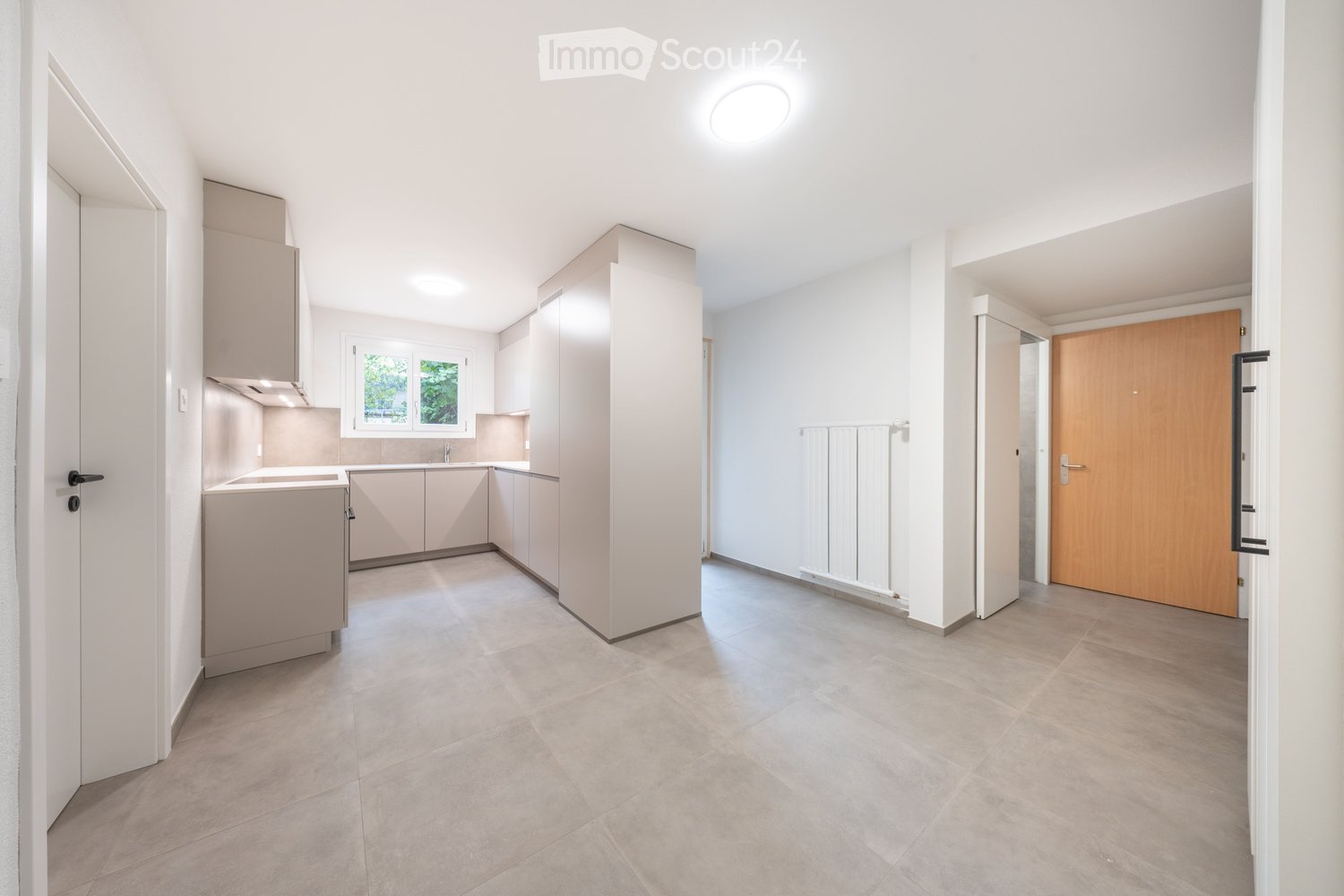 Empty kitchen and hallway area with light-colored cabinets, tiled floor, and a window. No furniture or appliances are present.