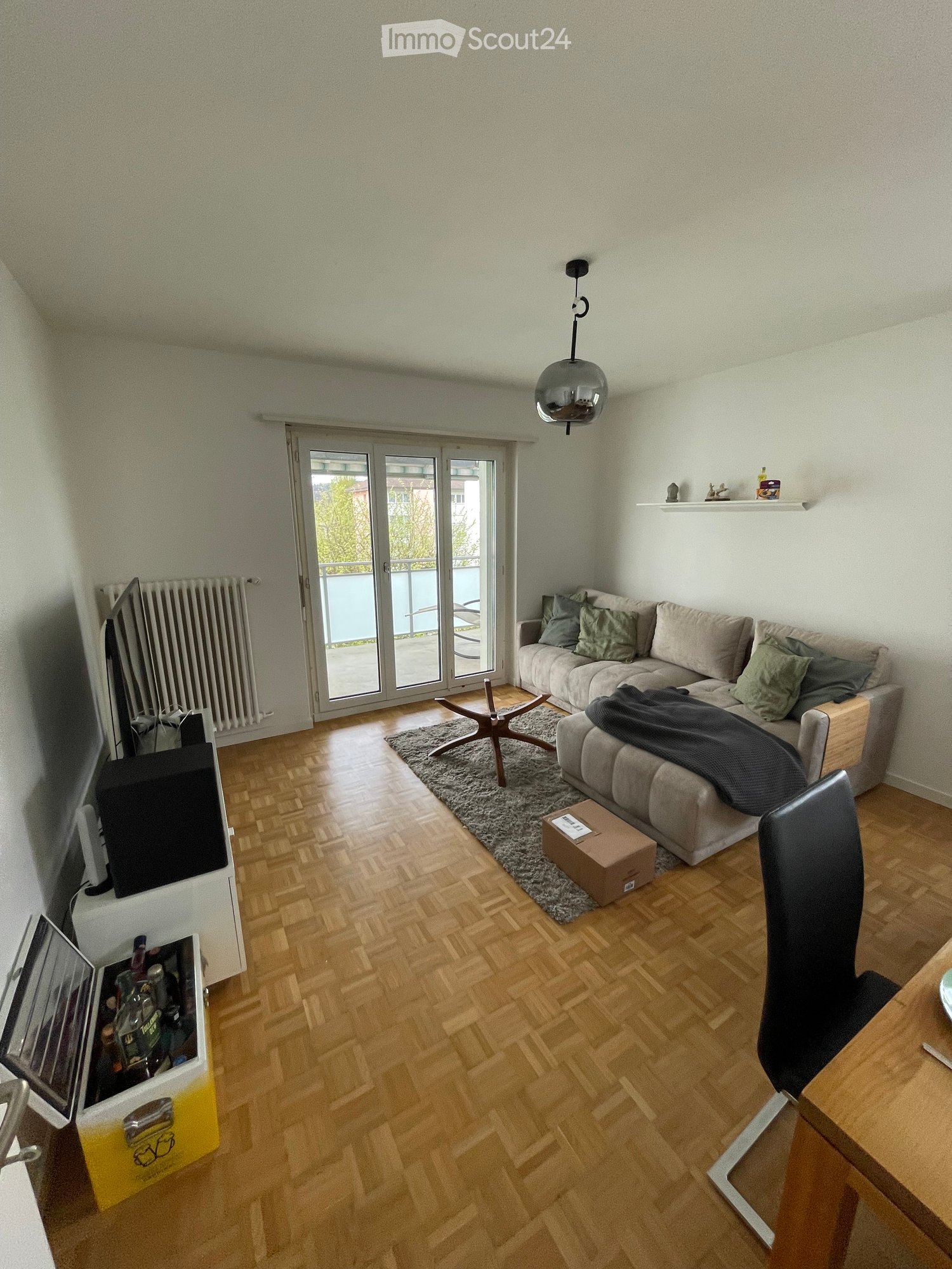 Living room with wooden flooring, a grey sofa with cushions, a coffee table, a hanging lamp, a television, and a sliding glass door leading to a balcony.