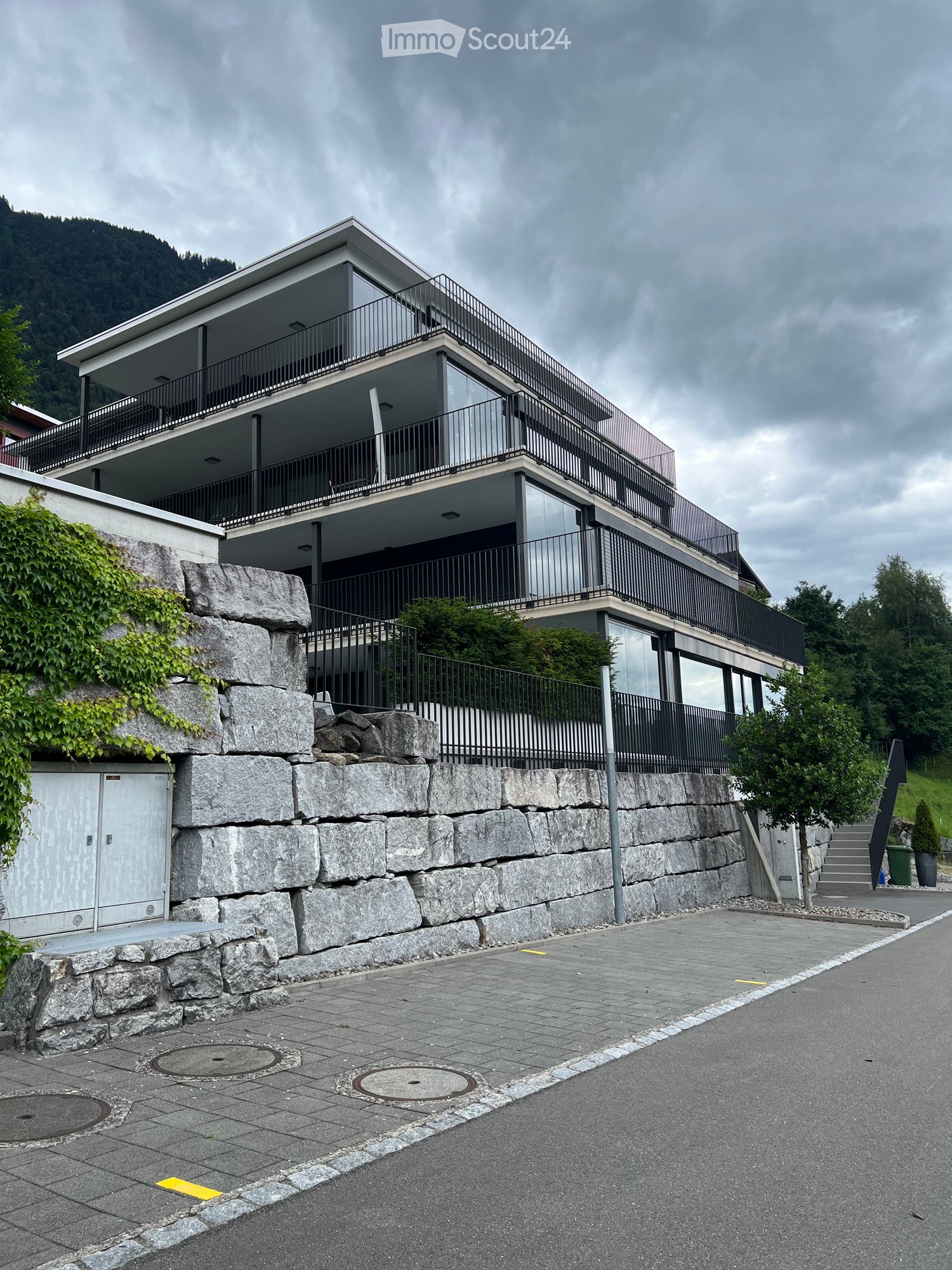 Modern two-story house with large windows and balconies, situated on a raised ground floor with stone retaining wall and greenery.