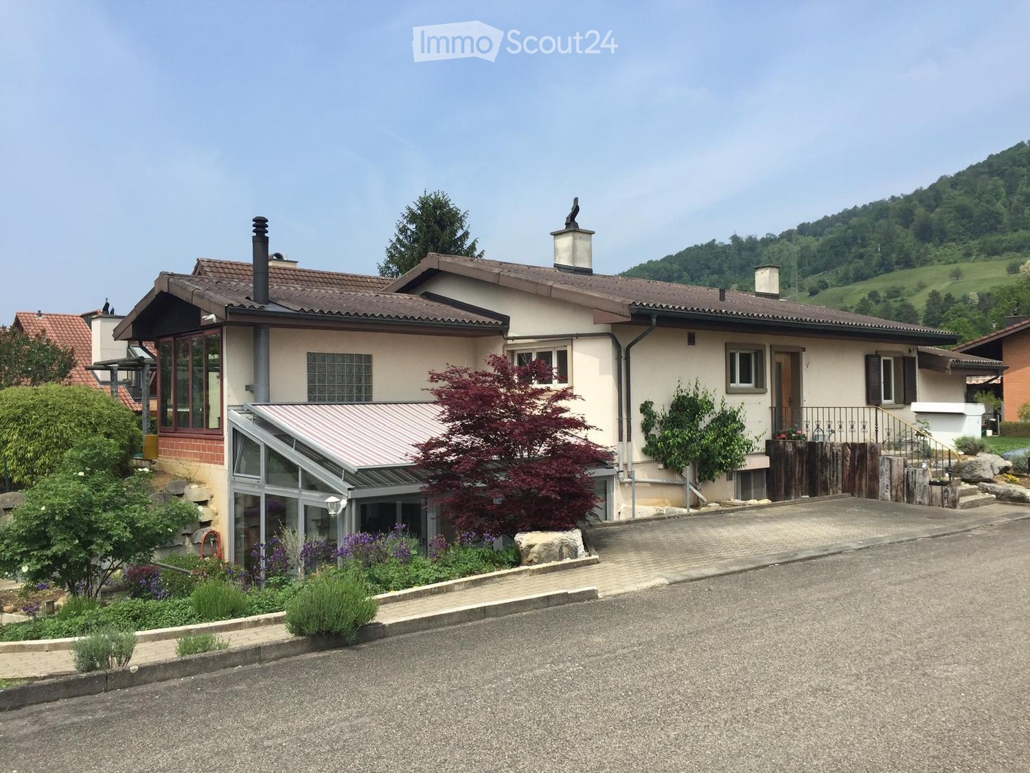 Two-story house with beige walls, red-tiled roof, multiple windows, and a small garden area with plants and a tree. The house features a covered entrance and is situated in a hilly, green landscape.