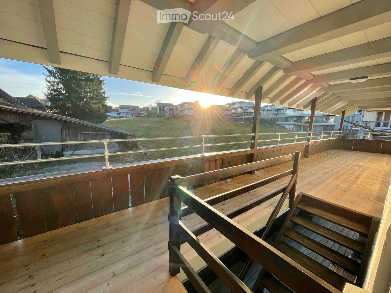 Wooden balcony with railing, steps leading down, and a view of a grassy area and neighboring houses.