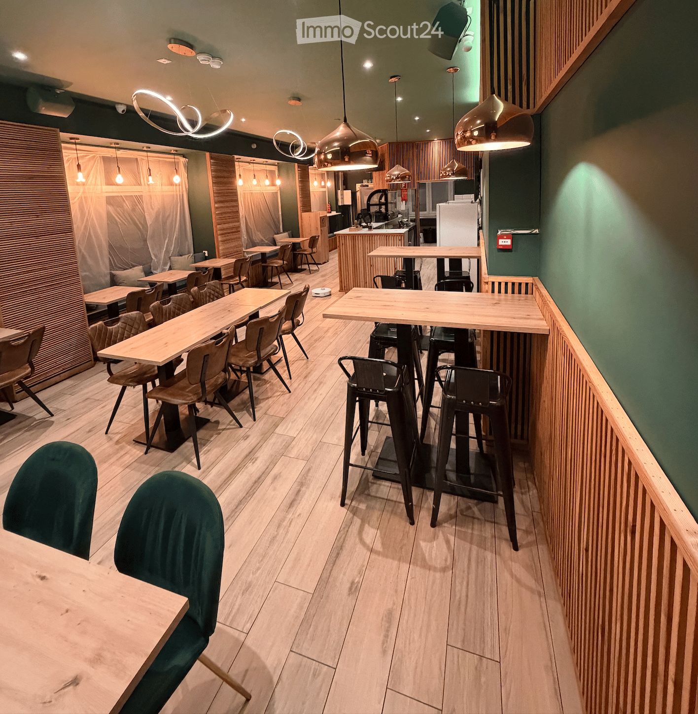 Modern restaurant interior with wooden tables, chairs, and stools arranged in a spacious layout. Green walls, pendant lighting, and a kitchen area visible in the background.