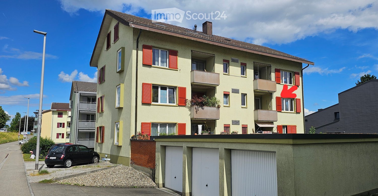 Multi-story residential building with beige facade, red shutters, balconies, and a parking area with three garage doors.