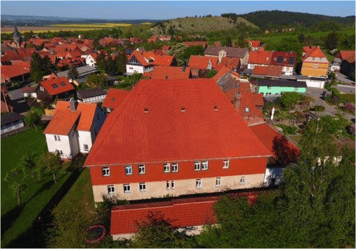 Aerial view of a large, red-roofed building surrounded by smaller houses and green fields in a rural village setting.
