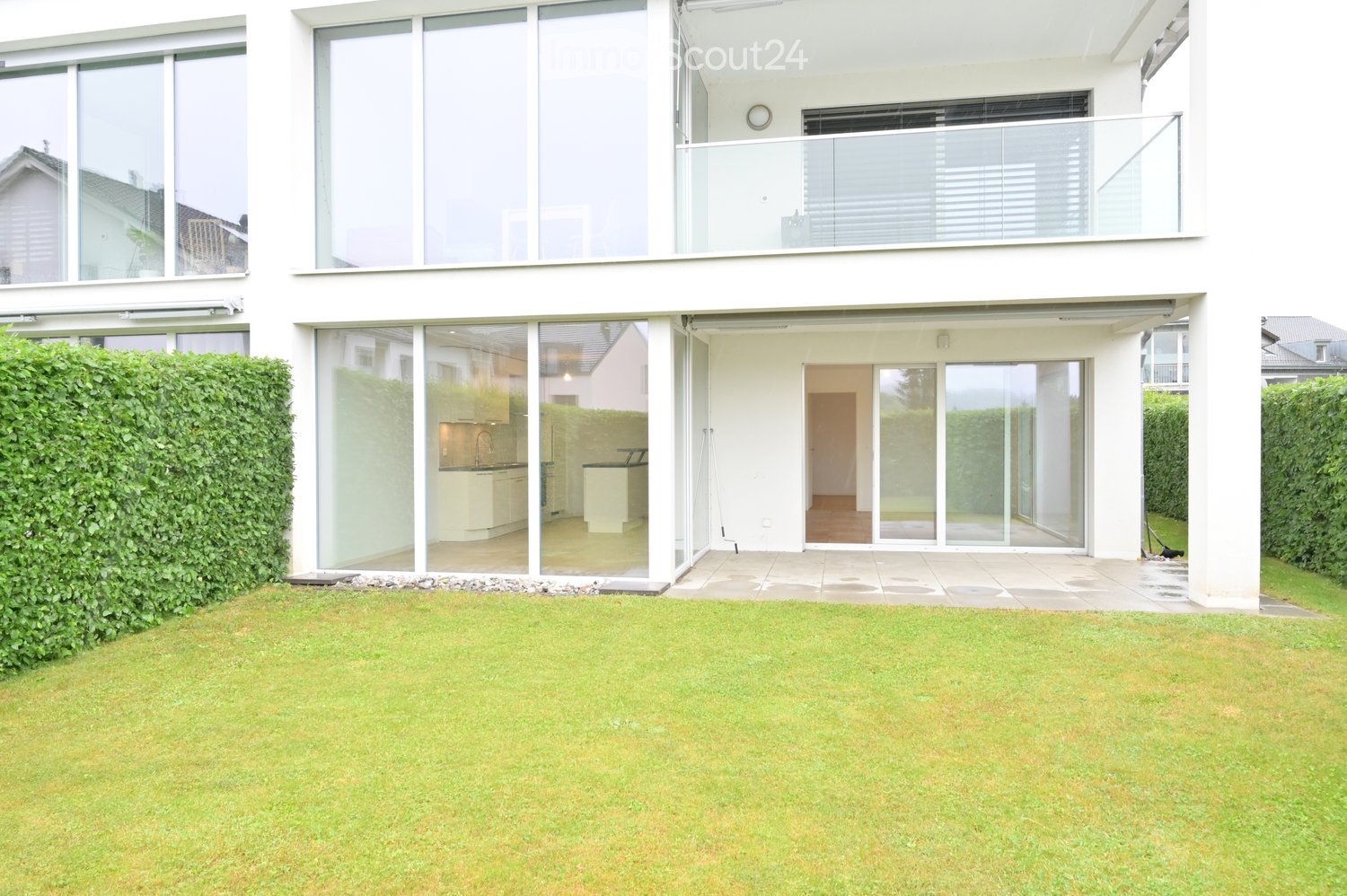 Modern two-story house with large glass windows and sliding doors, featuring a well-maintained lawn, neatly trimmed hedges, and a spacious patio area with a visible kitchen inside.