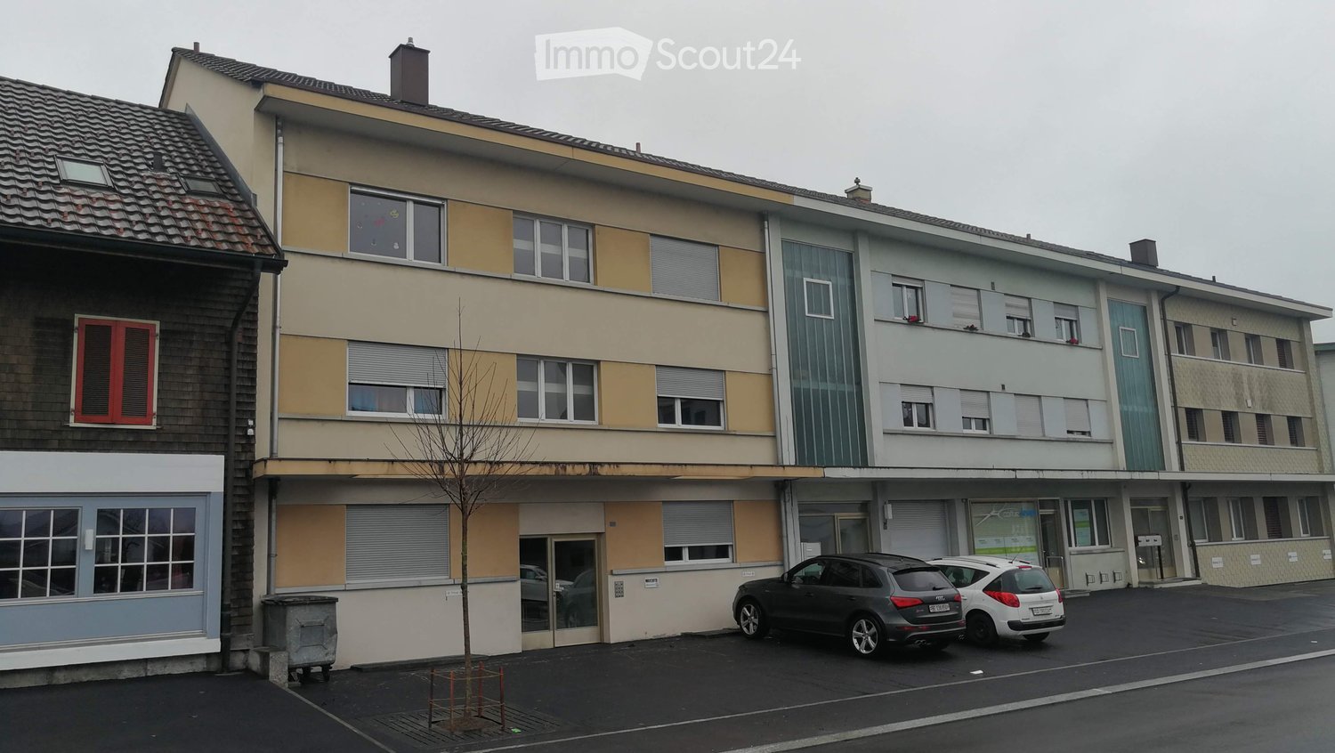 Multi-story residential building with varied facades, windows, and a small tree in front. Two cars are parked in front of the building.