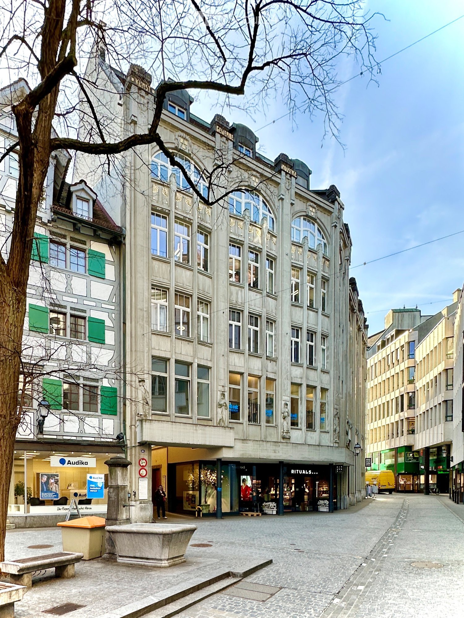 A multi-story building with ornate architectural details, large windows, and green shutters. The ground floor features storefronts, including a bank and a retail shop. There is a tree, a stone bench, and a trash bin in the foreground.