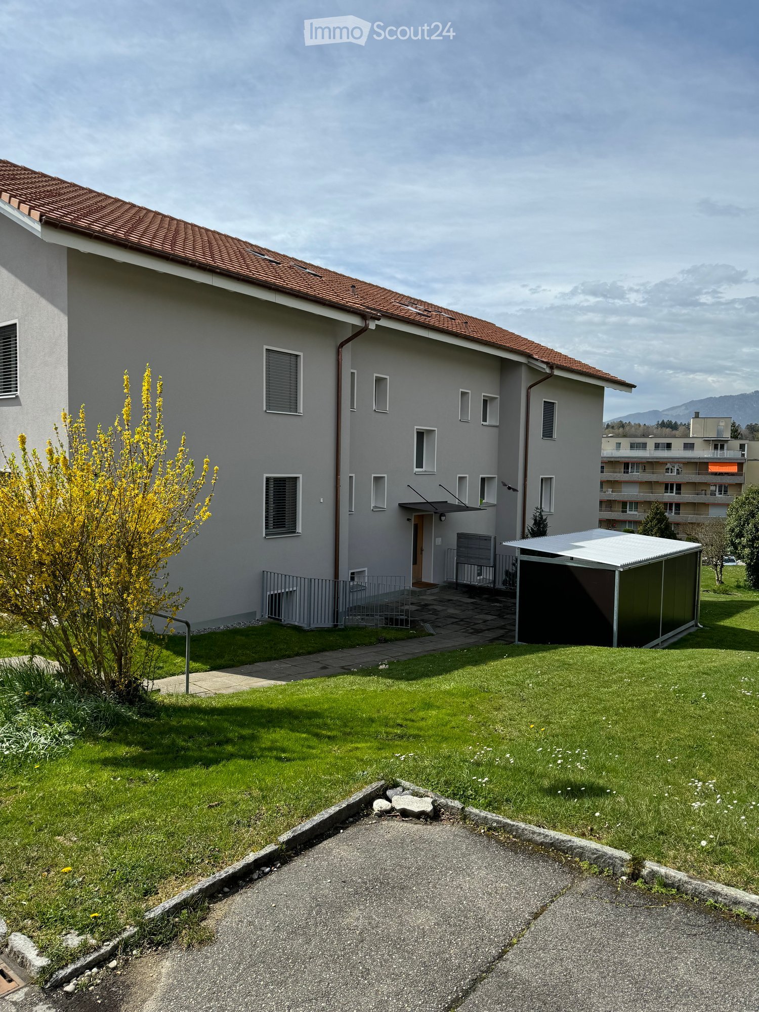 Two-story house with a red-tiled roof, grey exterior, multiple windows, and a small garden area with a pathway leading to the entrance.