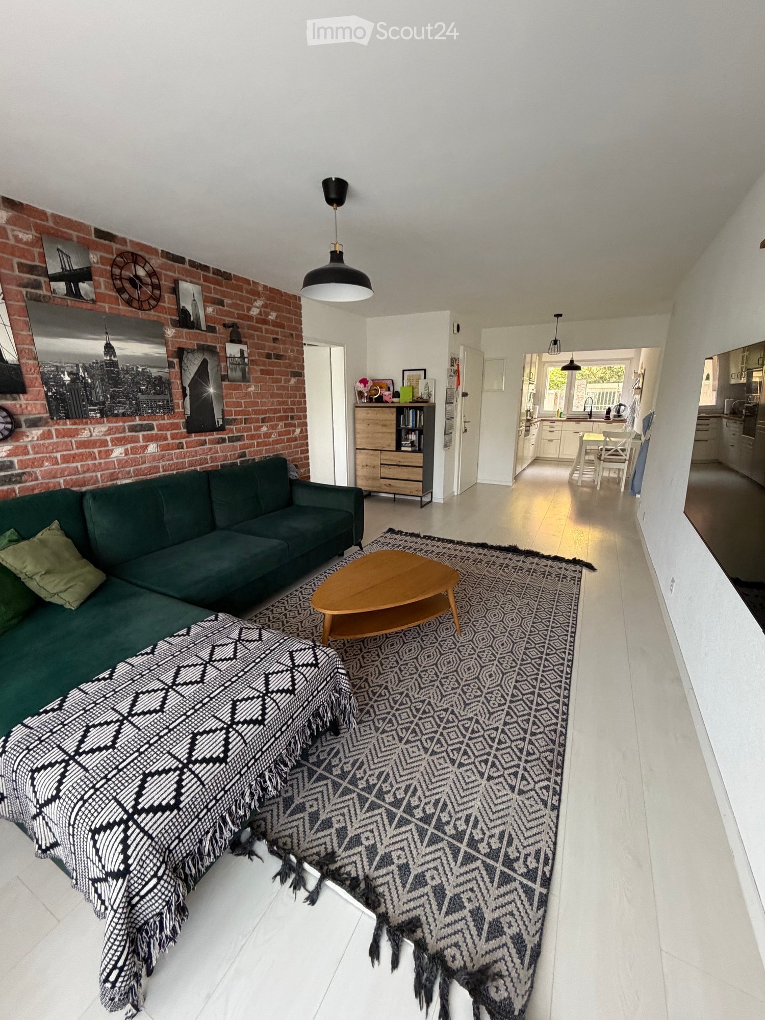 Modern living room with a green sofa, wooden coffee table, patterned rug, and exposed brick wall. The space is open-plan, connecting to a dining area and kitchen.