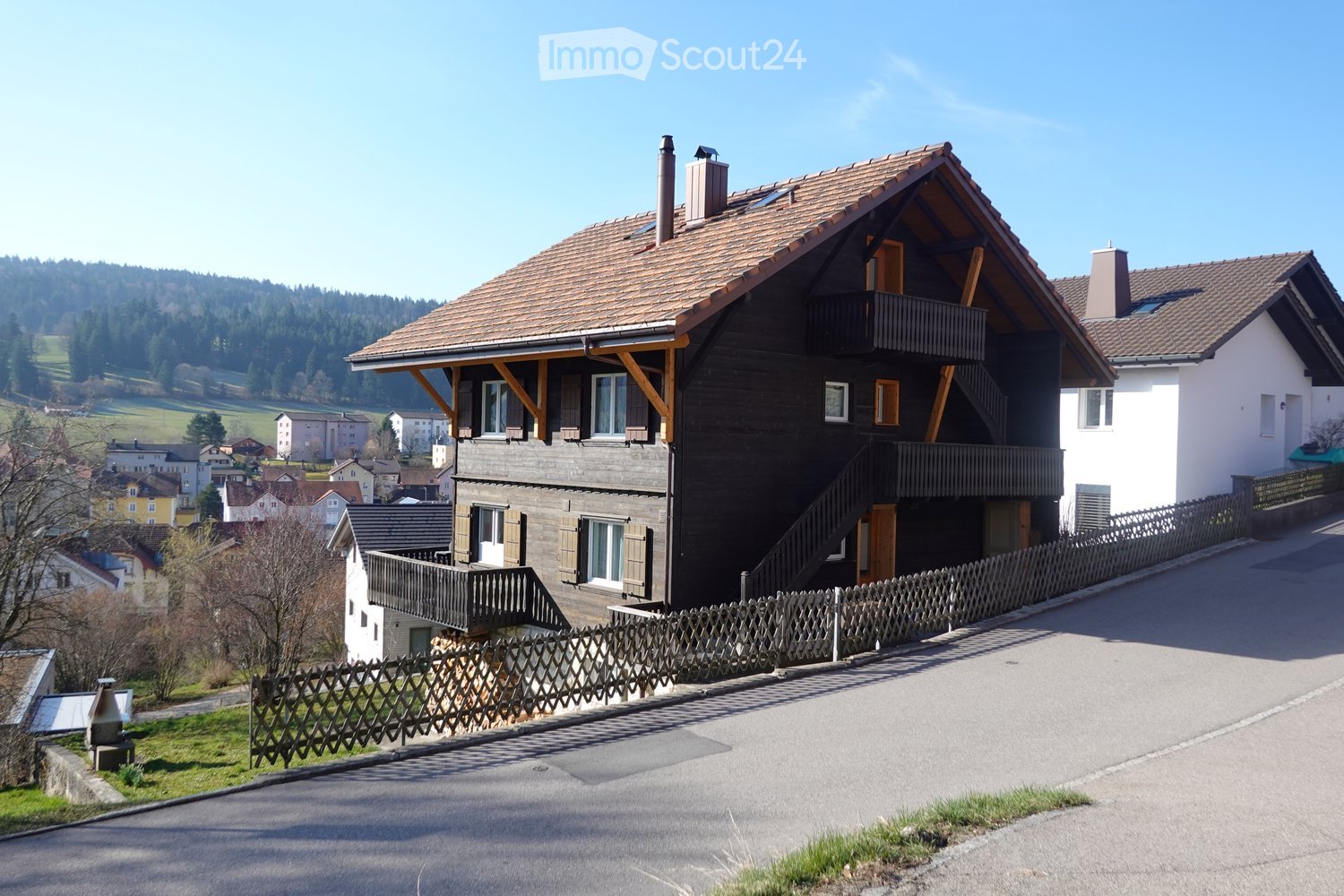 A two-story house with a dark wooden facade, a tiled roof, and a balcony overlooking a scenic valley.