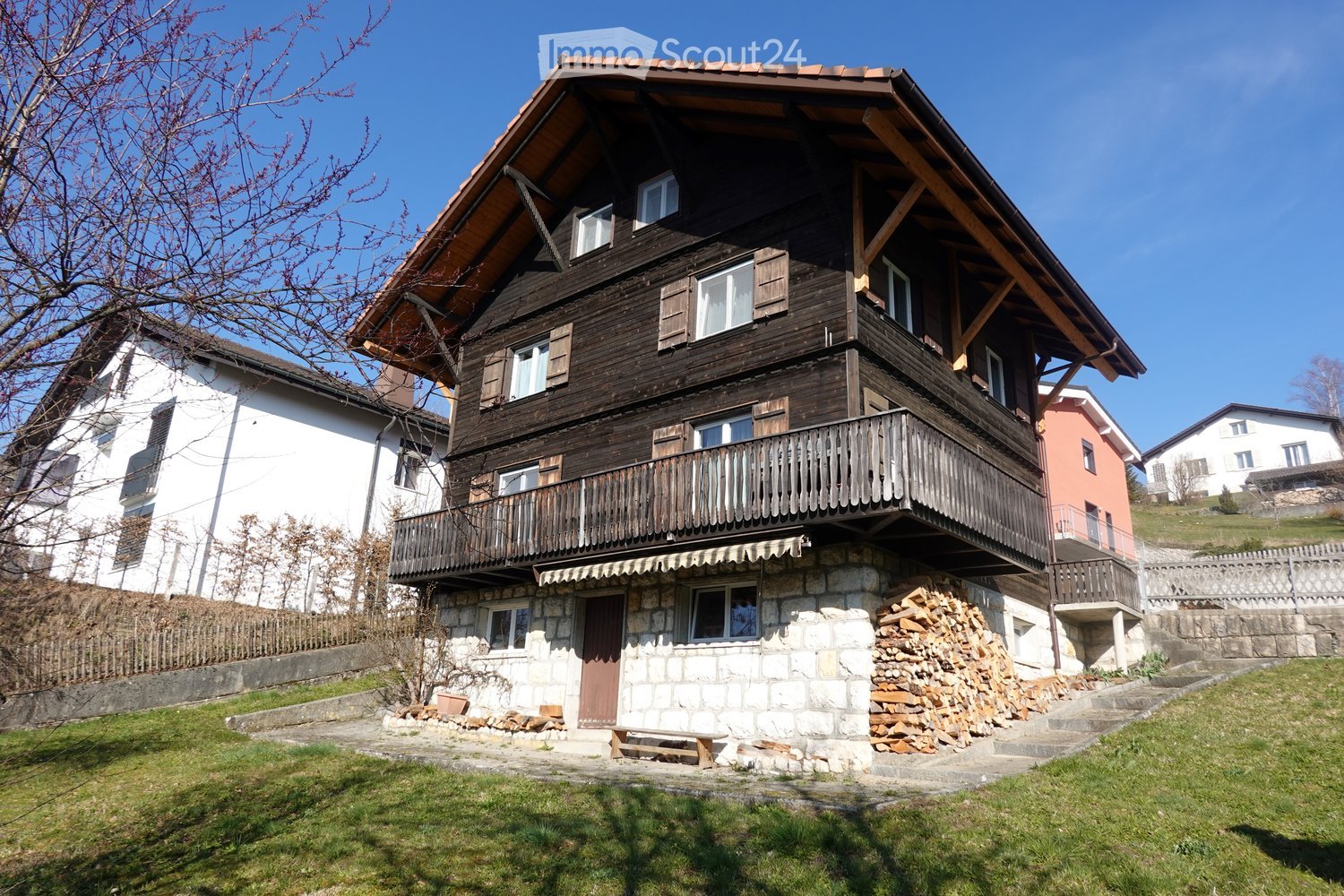 Two-story wooden house with a stone base, balcony, and wooden shutters. The house has a pitched roof and is surrounded by a grassy area with a stack of firewood and a bench.