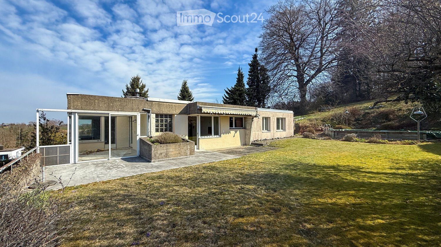 Modern single-story house with a flat roof, large windows, and a spacious lawn in front. The house features a concrete facade and a covered entrance area.