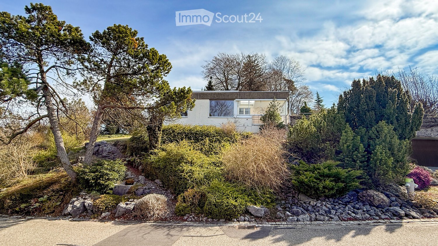A modern, single-story house with white walls and large windows, surrounded by lush greenery and trees. The house is elevated on a raised ground floor with a landscaped garden and rock formations in the foreground.