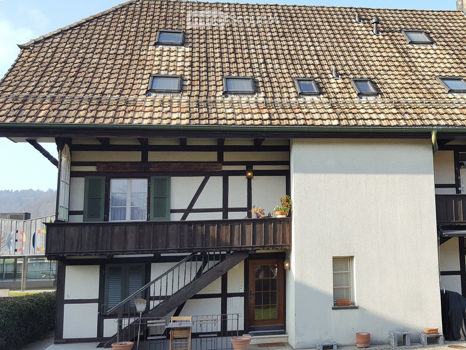 House. Traditional two-story house with a tiled roof, wooden balcony, and small garden area with potted plants and benches.