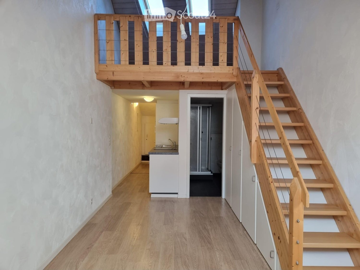 Empty room with wooden stairs, wooden floor, white walls, kitchen area with sink and shower