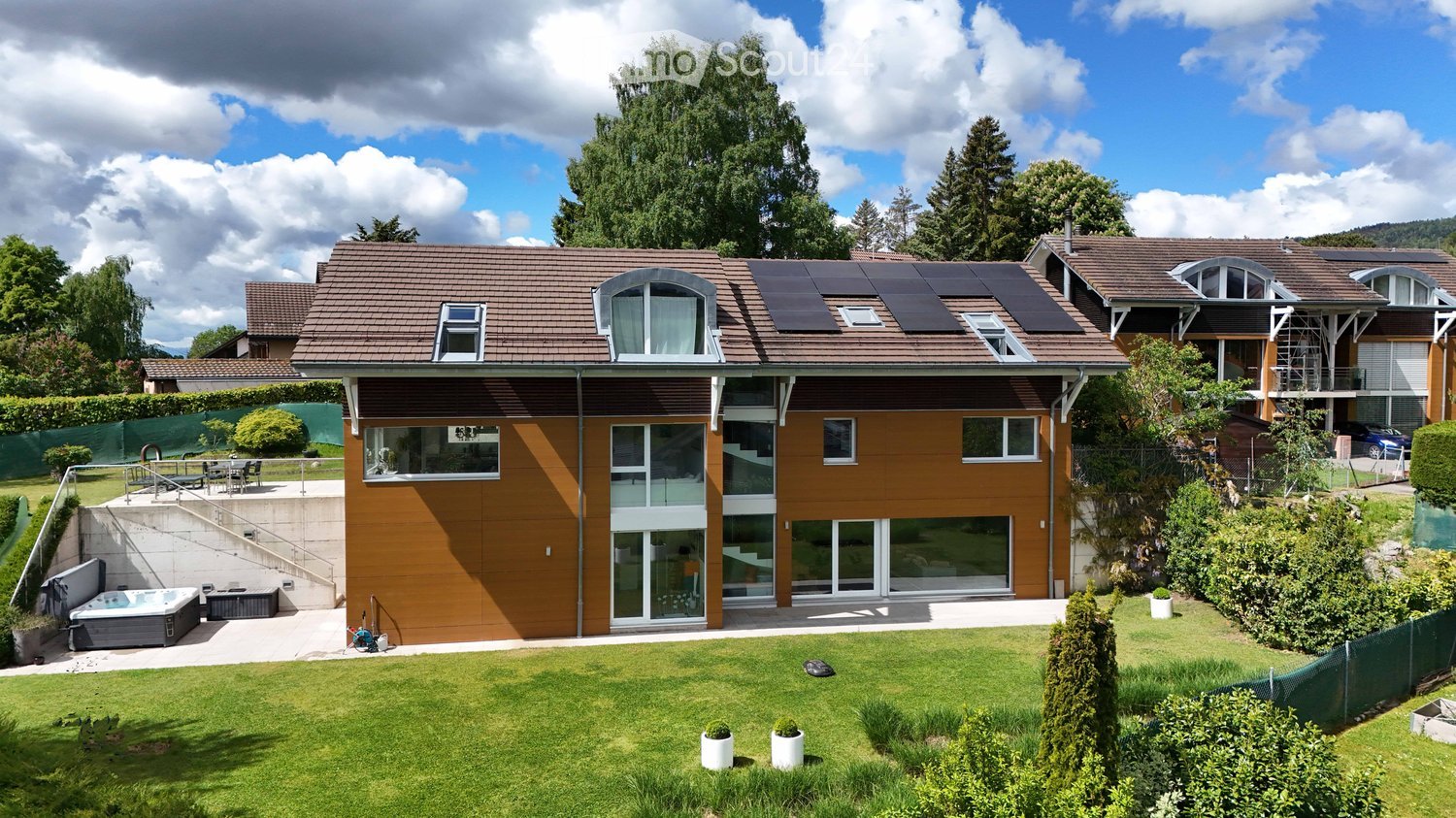 Modern two-story house with brown exterior, large windows, solar panels on the roof, and a well-maintained garden with a patio and hot tub.