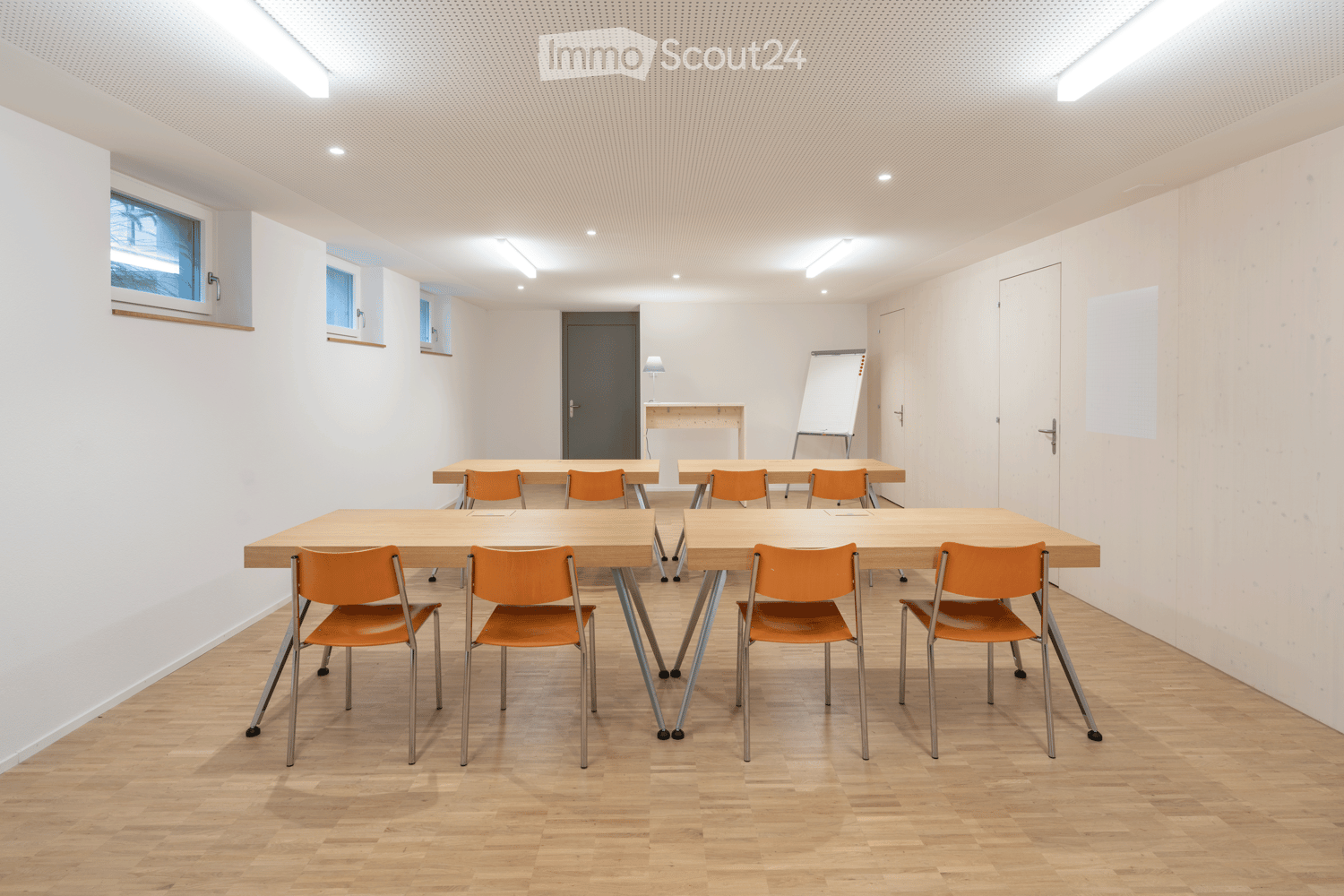 Bright, modern meeting room with light wood flooring, white walls, and multiple tables with orange chairs arranged for discussion.