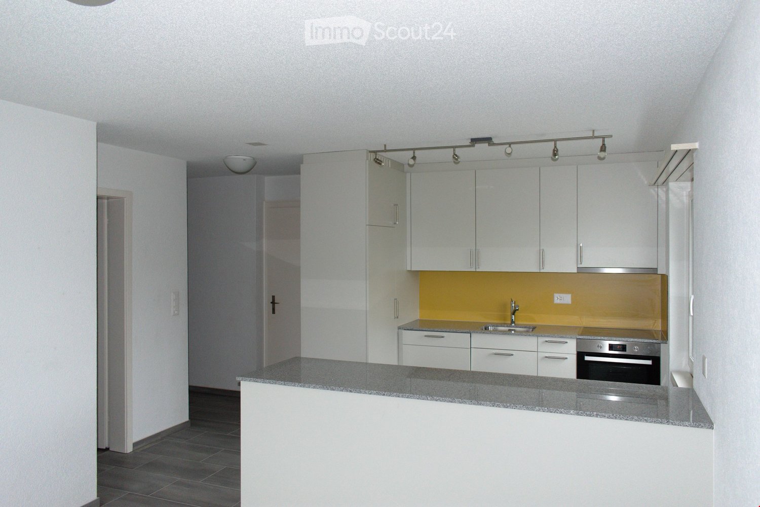 Modern kitchen with white cabinets, granite countertop, stainless steel sink, and built-in oven. Features a yellow backsplash and track lighting.