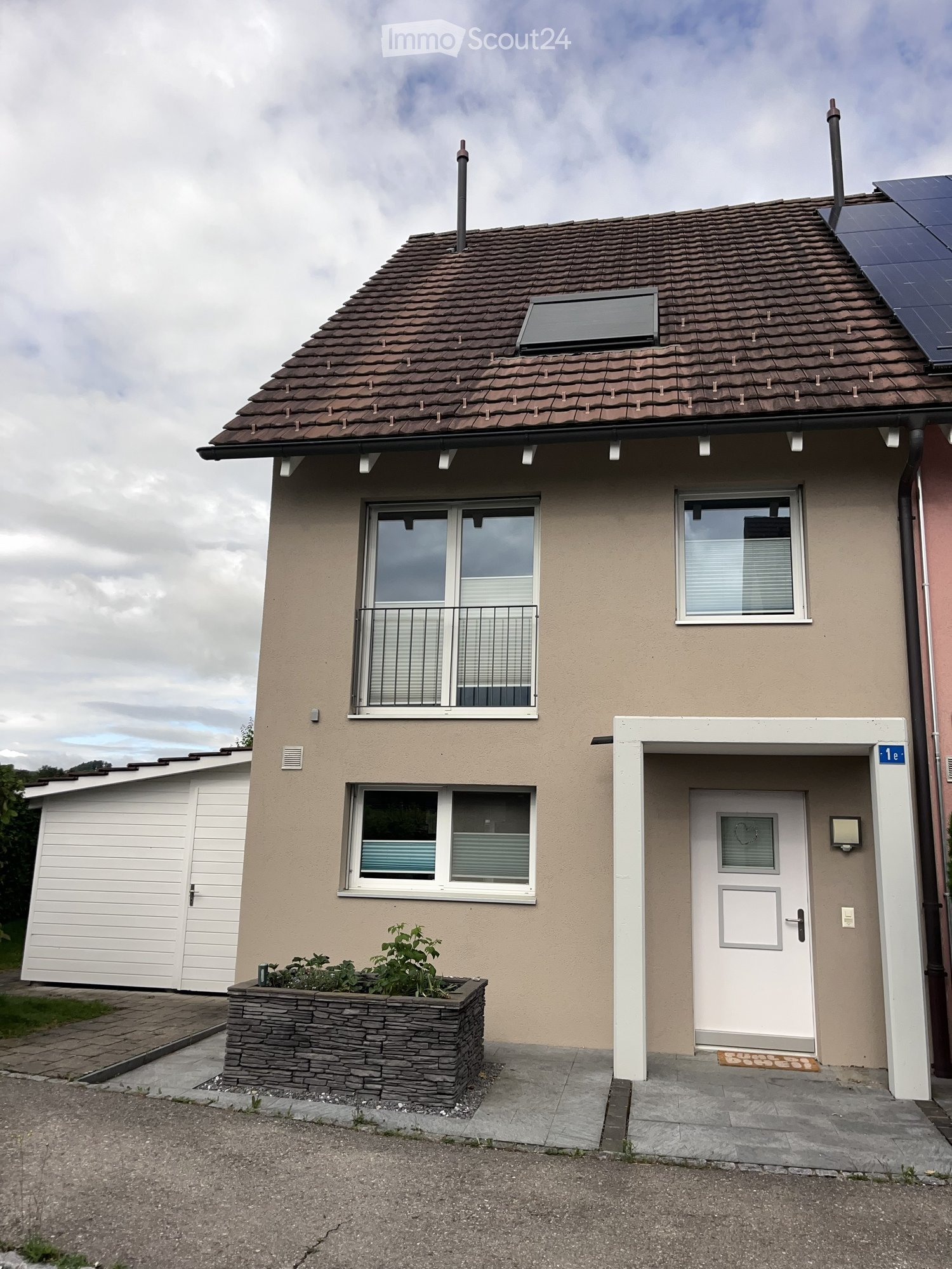 Two-story house with beige siding, tiled roof, white garage, and small balcony on the upper floor. Features include a front door with a decorative glass panel, a small planter, and solar panels on the roof.