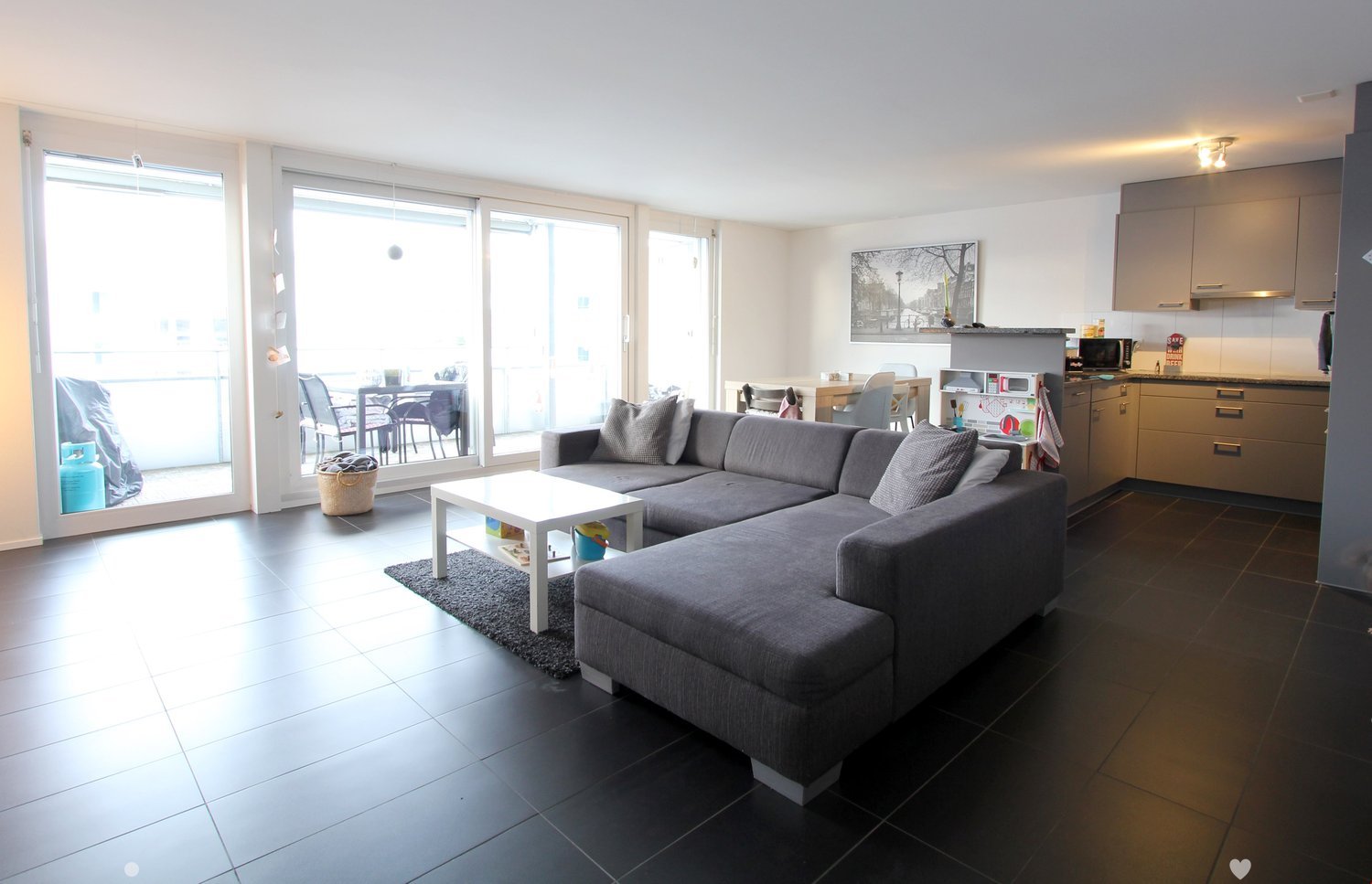 Modern living room with dark tiled flooring, grey sectional sofa, white coffee table, and a small dining area visible through large glass doors leading to a balcony.