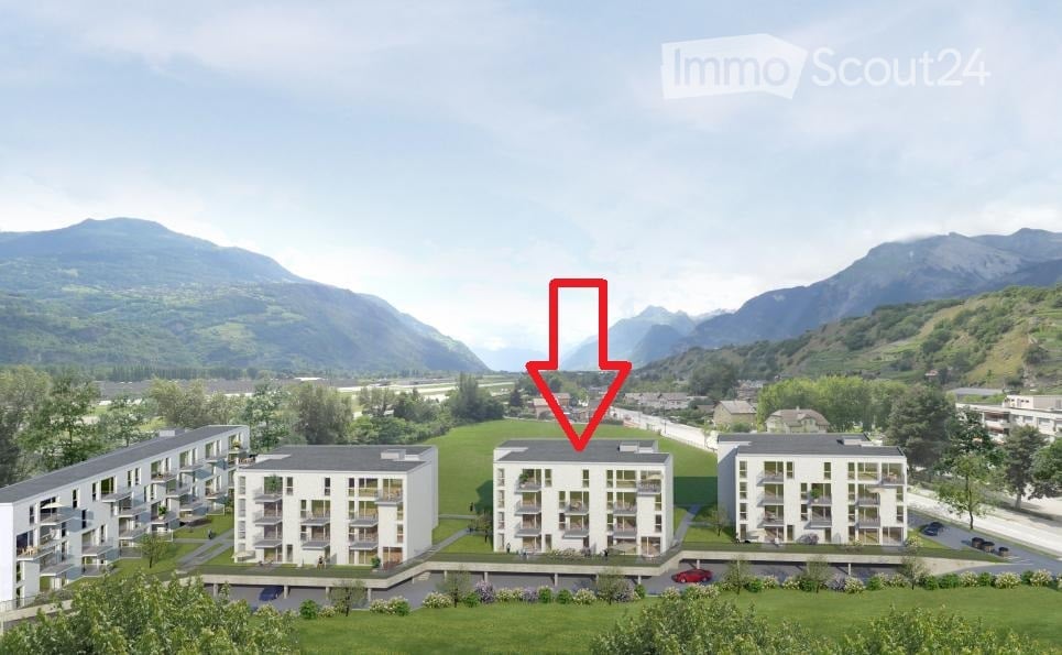 A modern multi-story residential building with balconies and a parking area, surrounded by green lawns and trees, with mountains in the background.