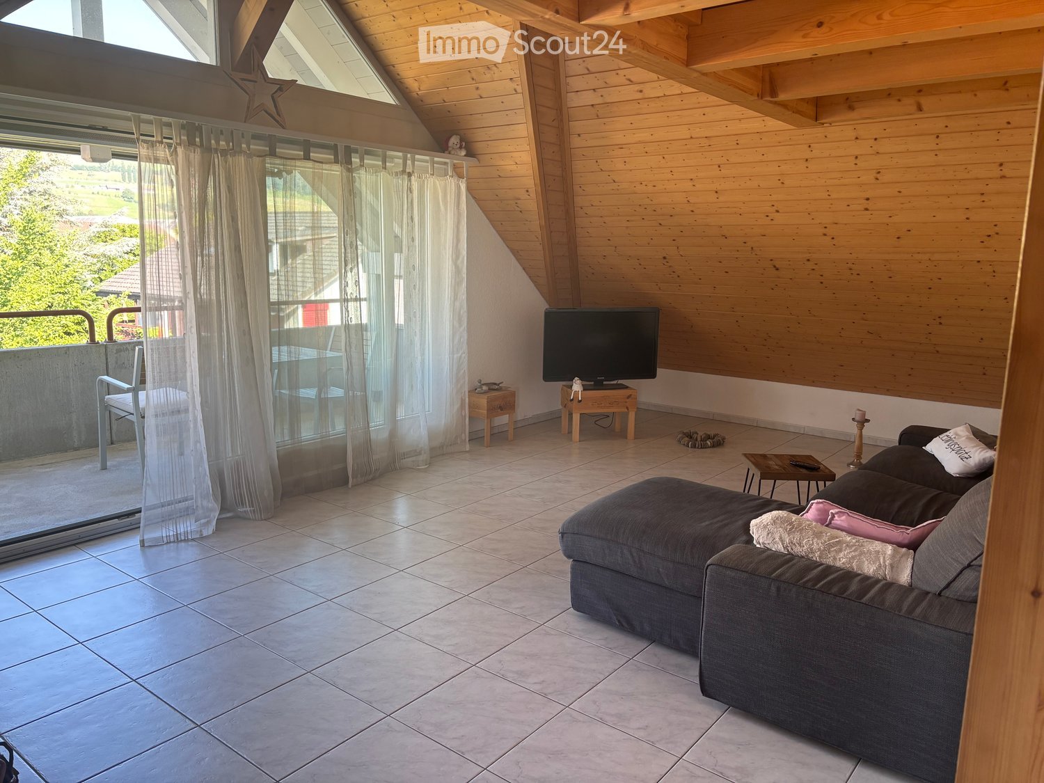 Modern living room with wooden walls, large windows, and a balcony. Features a grey sectional sofa, coffee table, TV, and a small table near the window.