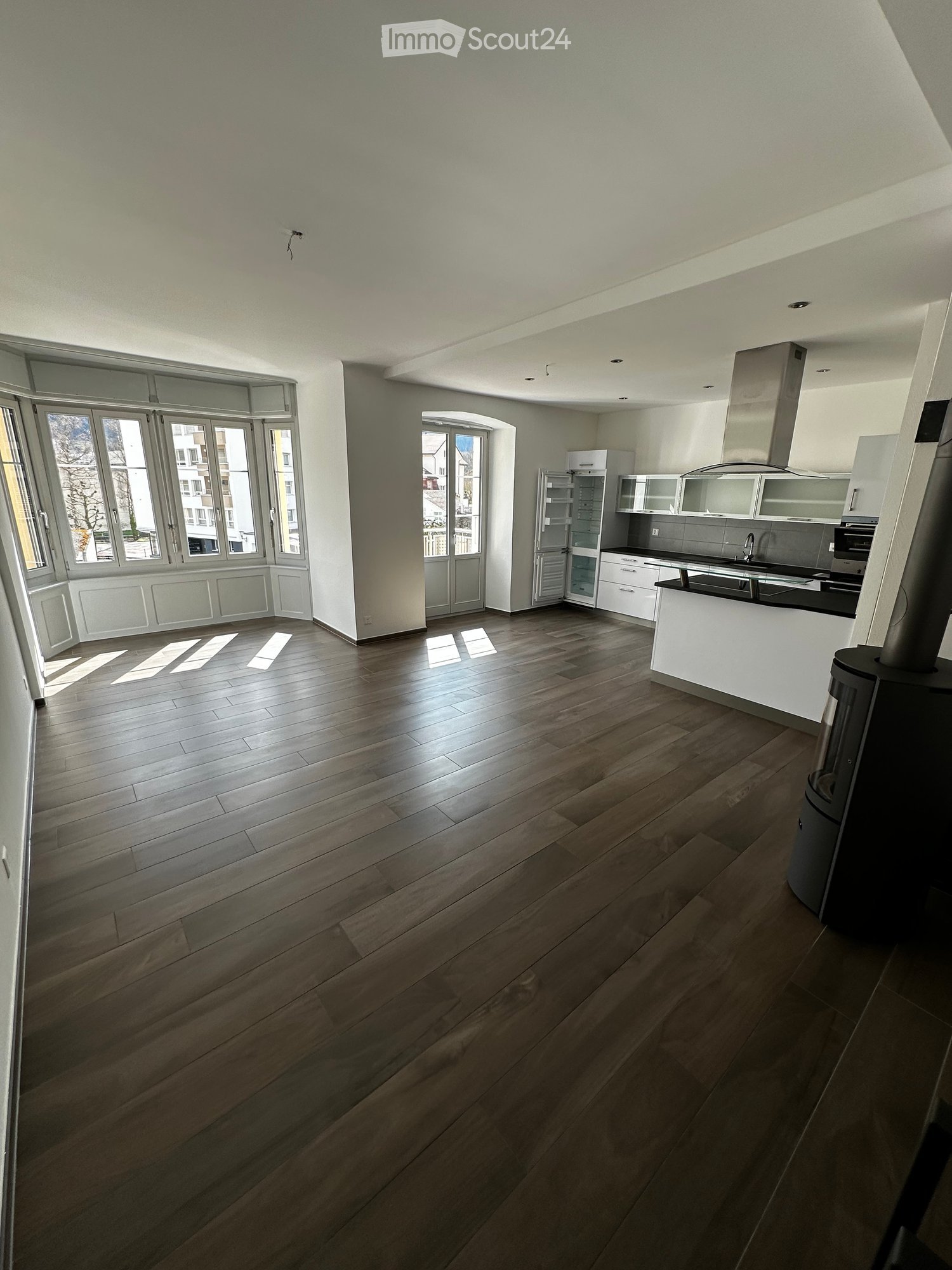 Open-plan space with wooden flooring, large windows, and a modern kitchen area featuring white cabinets, stainless steel appliances, and a central island.
