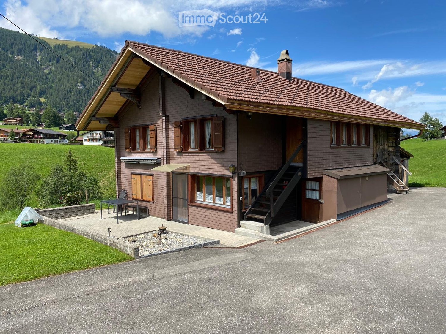 Two-story house with brown wooden exterior, red-tiled roof, multiple windows with shutters, and a small porch area with outdoor furniture.